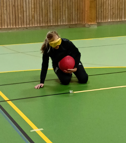 En person med långt ljust hår sitter på knä i en sporthall med grönt golv. Personen bär ögonbindel och håller i en röd boll, klädd i svarta träningskläder. Det är tydligt att hen deltar i en goalball-aktivitet. I bakgrunden syns en trävägg.
