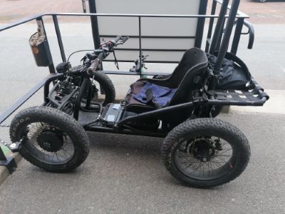 En svart, terränggående rullstol med fyra grova däck står parkerad på asfalt. Rullstolen har låg sittställning, fjädring framtill och handkontroller. Den är anpassad för att köras i ojämn terräng.
