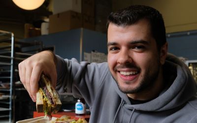 A young man with short dark hair and stubble smiles at the camera. He is holding up a large sandwich filled with various ingredients. He is wearing a grey hoodie and is sitting in what appears to be a kitchen or bakery, with shelves and boxes in the background.