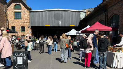 People walk and gather between market stalls with colorful tents. In the background, a large brick industrial building is visible.