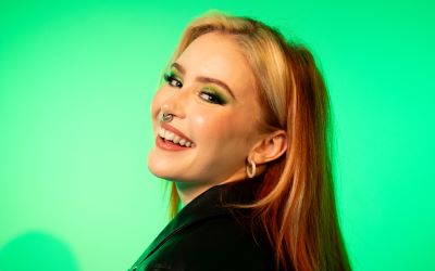 A woman is smiling broadly in front of a green background. She has orange-colored hair and a rock-inspired makeup look, with green eyeshadow and a septum piercing.