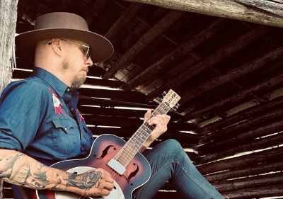 A man with tattoos, a hat, sunglasses, and a country shirt is seen in profile, playing the guitar. He is in a setting that resembles an old barn.mmal lada.