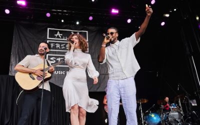 A stage scene from a concert. Three performers stand at the front: one man playing an acoustic guitar, a woman in a white dress singing into a microphone, and another man in white clothes rapping or singing with one arm raised. In the background, a drummer is visible. Behind them hangs a black backdrop with the text “All My Friends Are Stars Music Festival”.