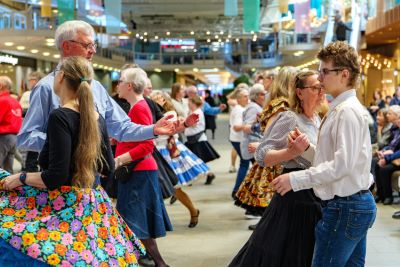 Ett dussin människor, kvinnorna iklädda långa, färgglada kjolar och männen bär skjorta. De dansar squaredans i par. Dansen äger rum i ett köpcentrum.