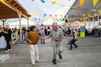 Två pensionärer dansar bugg på en träterrass. Terrassen är dekorerad med färgglada flaggor och runt om de syns andra dansande par och människor som samtalar och minglar.
