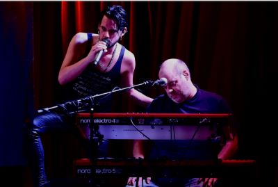 A stage scene with two musicians. On the left, a man leans against a microphone stand, singing into a microphone. He is wearing a sparkling black tank top and black pants. On the right, a man is seated, playing two red electric keyboards (Nord Electro). He is dressed in a dark t-shirt and has a shaved head. A dark red curtain is visible in the background.