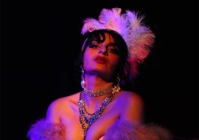 En person sitter på en stol i scenljus med blått och rött sken. Hen är klädd i burlesk stil med fjäderboa, nätstrumpbyxor, smycken och en huvudprydnad med fjädrar. Personen lutar sig lätt framåt och ser uppåt med ett uttrycksfullt ansiktsuttryck.